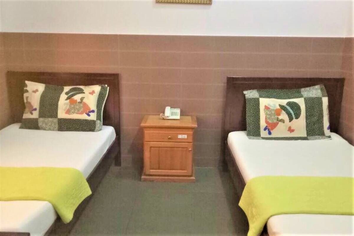 A hotel room with two beds, each with a quilted headboard and a yellow blanket. There is a wooden nightstand with a telephone between the beds.