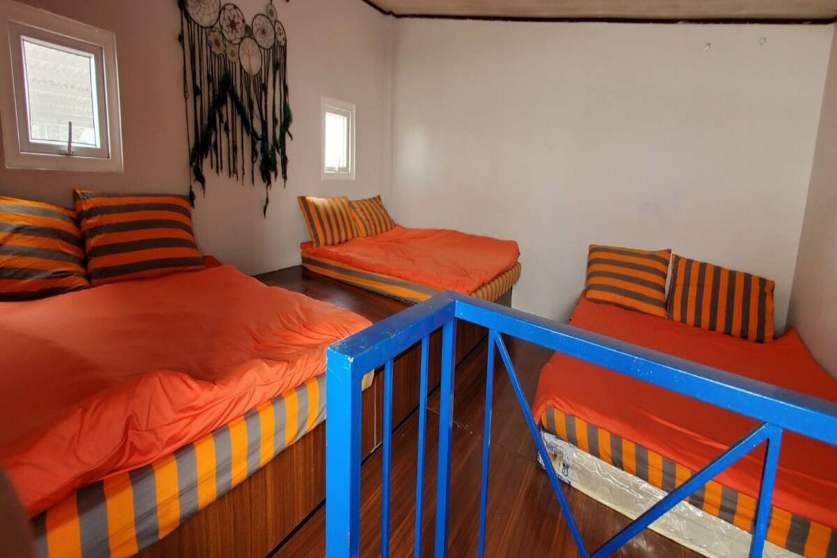 A room with three beds, each with orange bedding and striped pillows. There are two windows and a dreamcatcher on the wall.