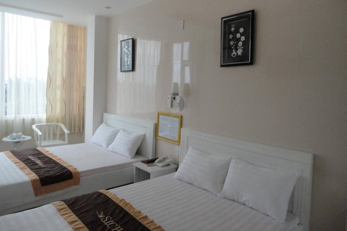 A hotel room with two beds, a small table, a chair, and a window with curtains.