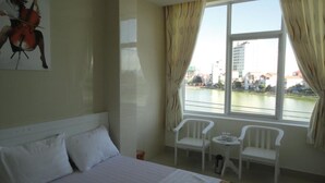 A hotel room with a bed, two white chairs, a small table, and a view of a river and buildings. - SIC Hotel (Dong Hoi)