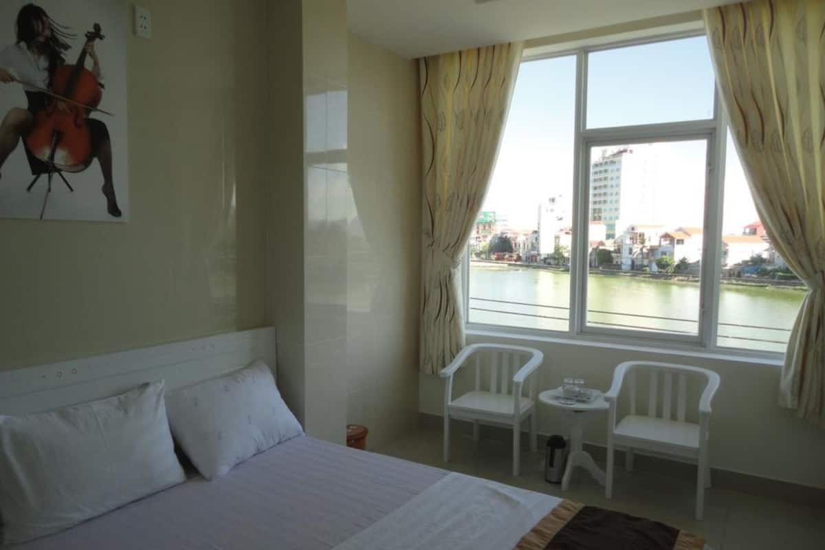 A hotel room with a bed, two white chairs, a small table, and a view of a river and buildings.