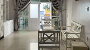 Uma sala de estar espaçosa com ventilador de teto, um sofá branco, uma cadeira de madeira e um banco. Há uma porta de vidro que dá para o exterior, e um armário branco é visível no canto.