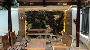 Uma área de estar coberta ao ar livre, com um muro de pedra ao fundo, mobiliário de madeira e piso estampado.