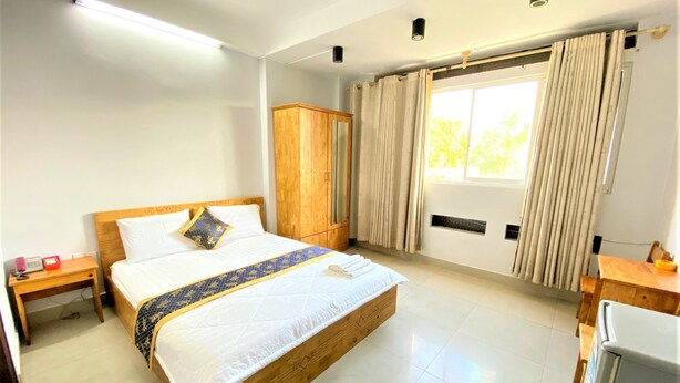 A hotel room with a large bed, wooden headboard, a wooden wardrobe, a desk with a chair, a small table, and a window with curtains. - Thien Son Hotel (Ho Chi Minh City)
