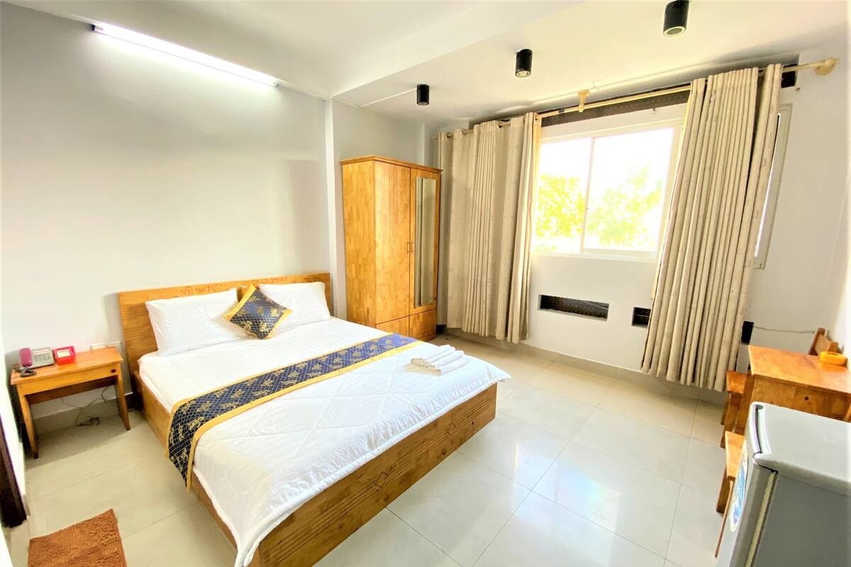 A hotel room with a large bed, wooden headboard, a wooden wardrobe, a desk with a chair, a small table, and a window with curtains.