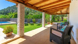 An outdoor patio with a pergola, stone pillars, wicker furniture, and a view of a pool and greenery.