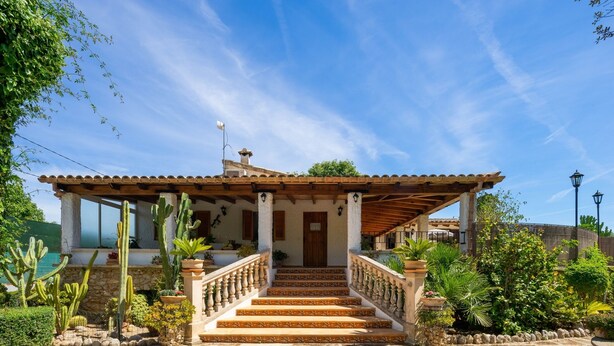 A two-story house with a wooden porch, stone steps, and a garden with cacti and plants.
