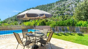 A poolside area with a table and chairs, a covered umbrella, and a view of a mountainous landscape.