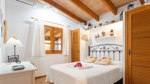 A bedroom with a bed, a bedside table, a lamp, a window with curtains, and a shelf with decorative items.