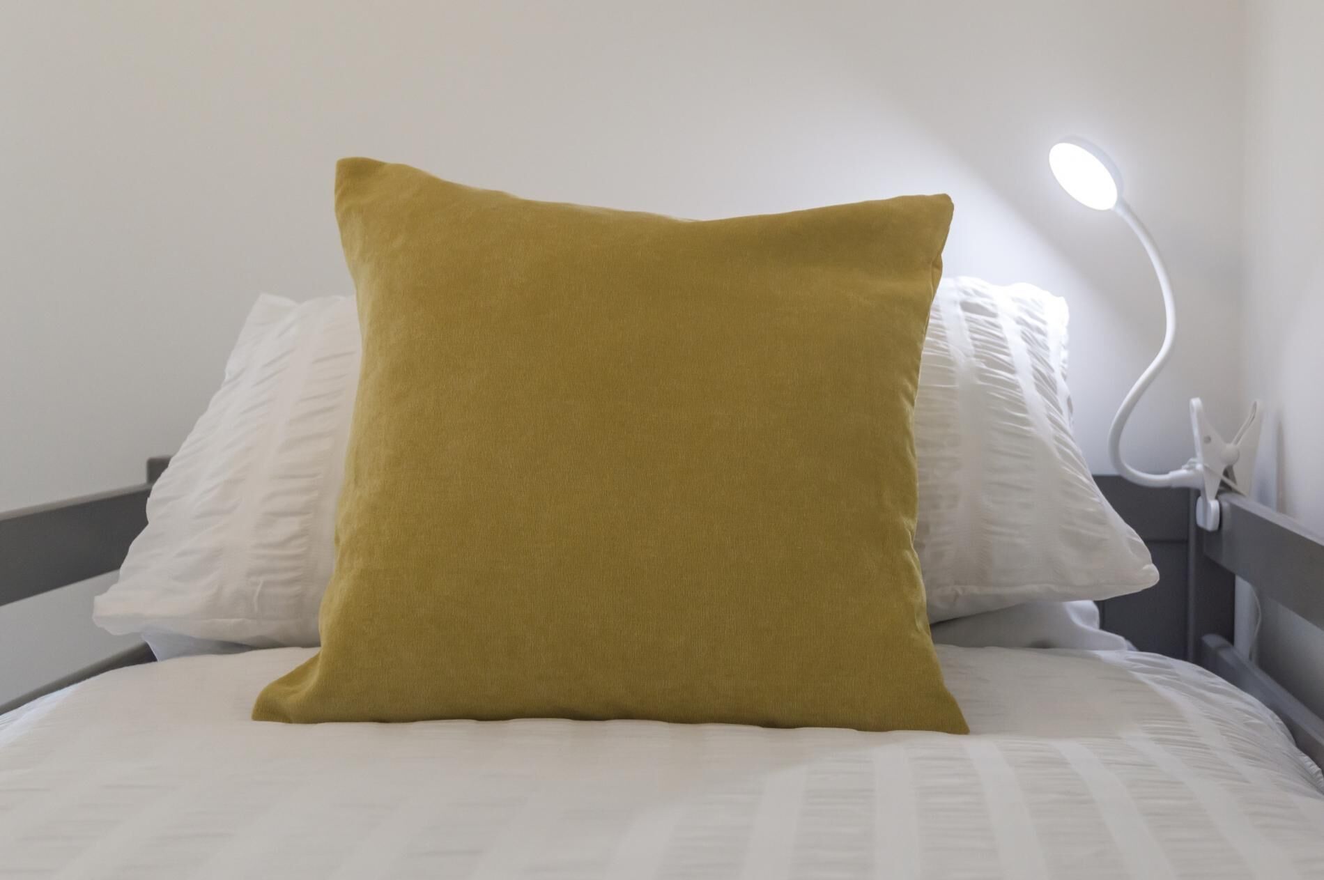 A bed with a single mustard-colored pillow and a white pillow, a white nightstand with a lamp, and a plain wall.