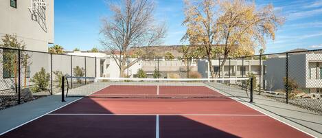 An outdoor tennis court with a net, surrounded by a fence, and trees in the background.