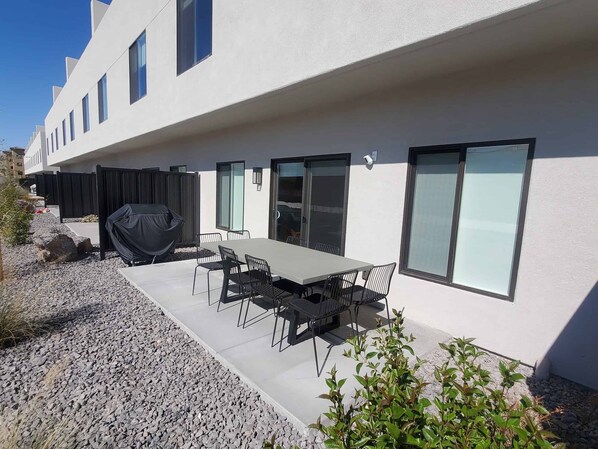 A modern outdoor patio with a dining table and chairs, a barbecue grill, and a white building with large windows.