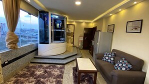 A modern living room with a curved entertainment unit, a sofa with patterned cushions, a small table, and a view of the city through large windows. - Deluxe Studio with amazing Pyramids view (Giza)