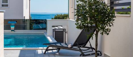Un espace piscine moderne offrant une vue dégagée sur la mer, avec un fauteuil de relaxation et un petit arbre.