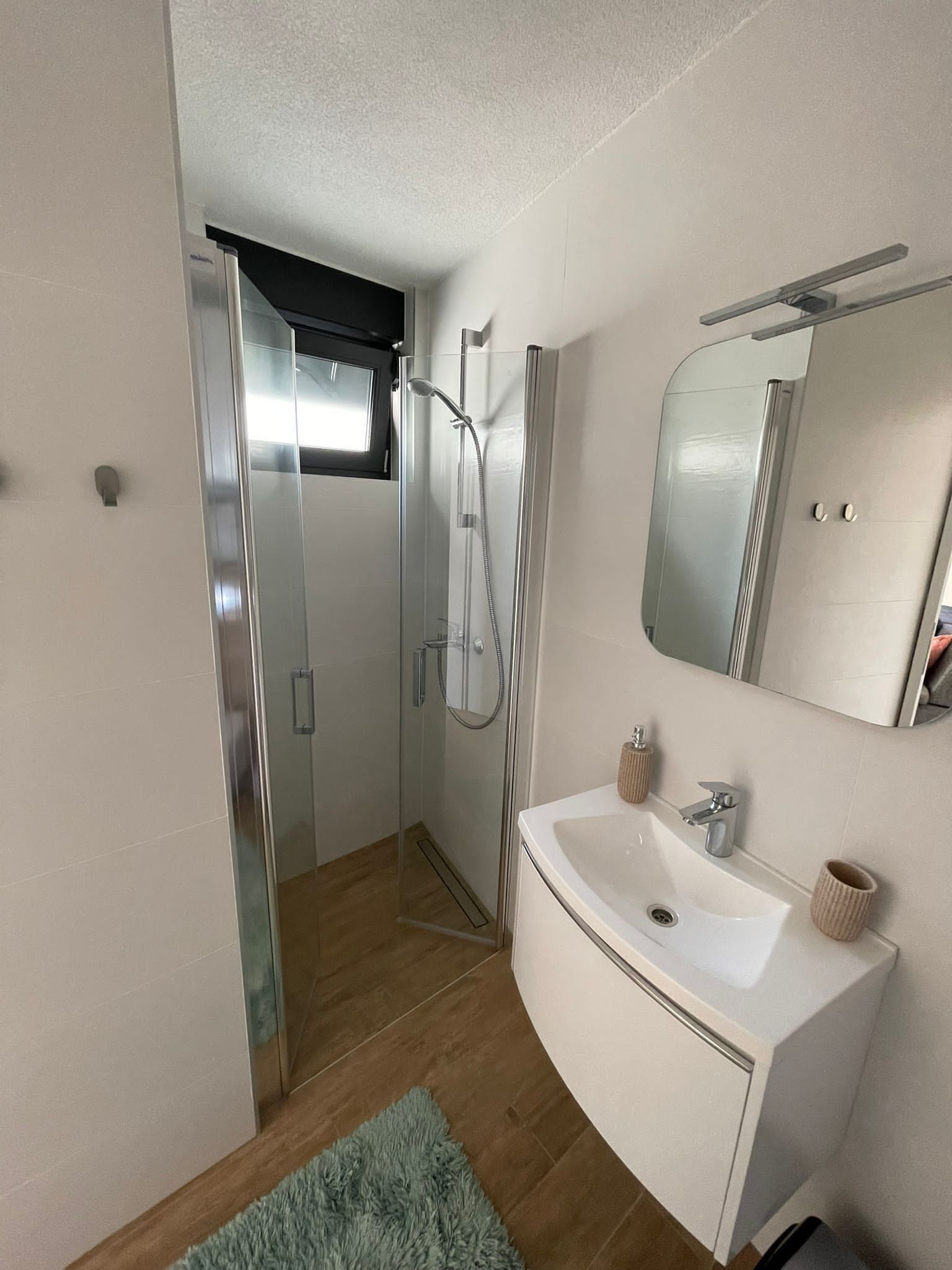 A modern bathroom with a glass-enclosed shower, a white sink with a mirror, and a fluffy blue rug on the floor.