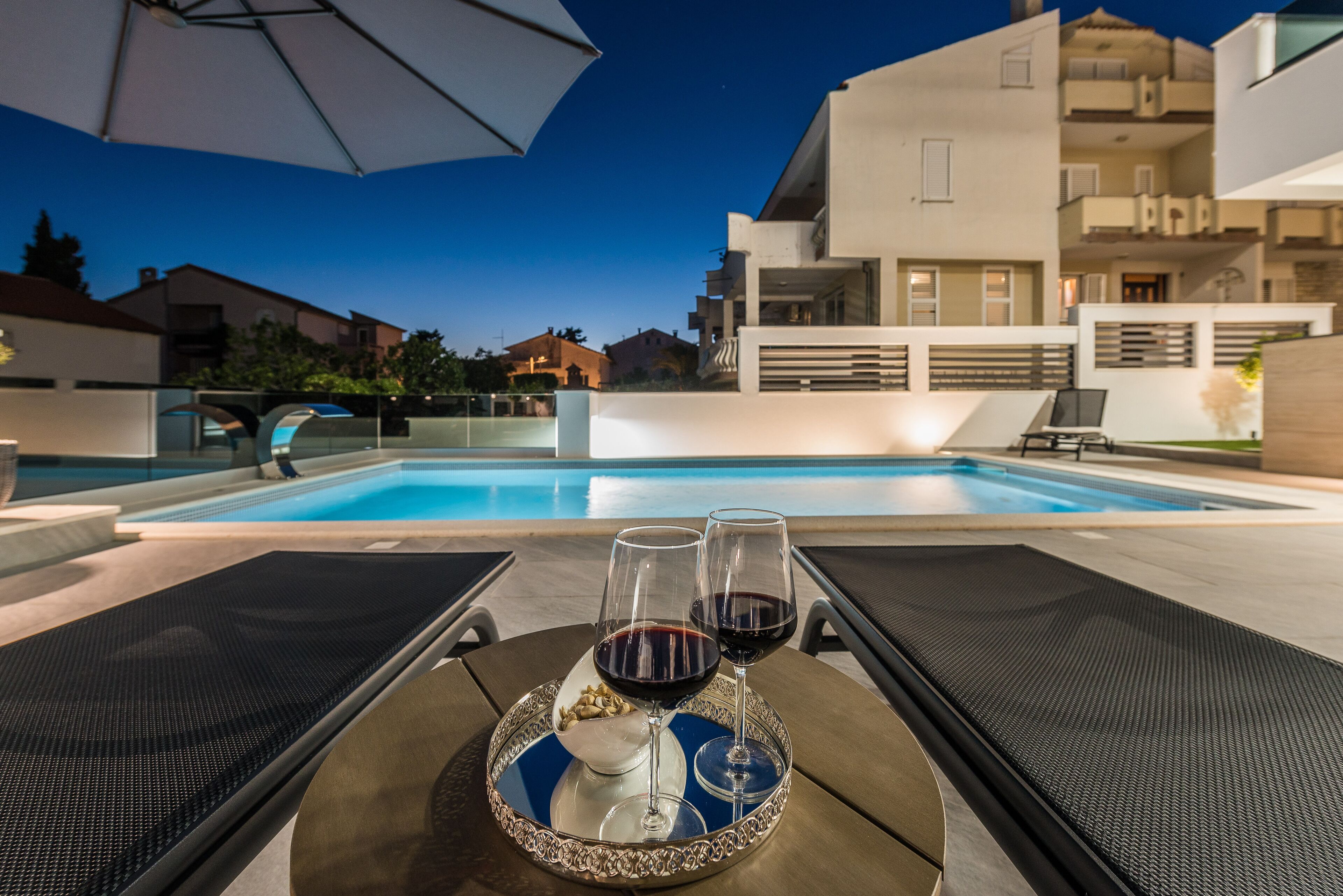 Vista de la piscina con dos sillas de descanso, una bandeja con dos copas de vino y un bol de frutos secos, y un edificio moderno al fondo.