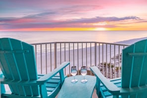 A balcony with turquoise chairs and a table, overlooking a beach with a sunset view.