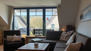 A cozy living room with a brown sofa, a wooden coffee table, and a chair. A large window offers a view of a balcony and a building outside. - Papillon Wohnung 08-7 (Ostseebad Boltenhagen)