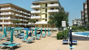 Un hotel fronte spiaggia con numerosi balconi, una zona piscina con lettini e ombrelloni, e un cielo sereno.
