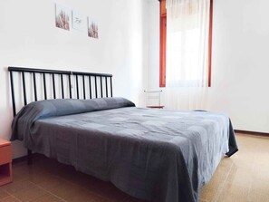 A bedroom with a bed, a nightstand, a chair, and a window with curtains. - Studio directly on the beach (Lido di Jesolo)