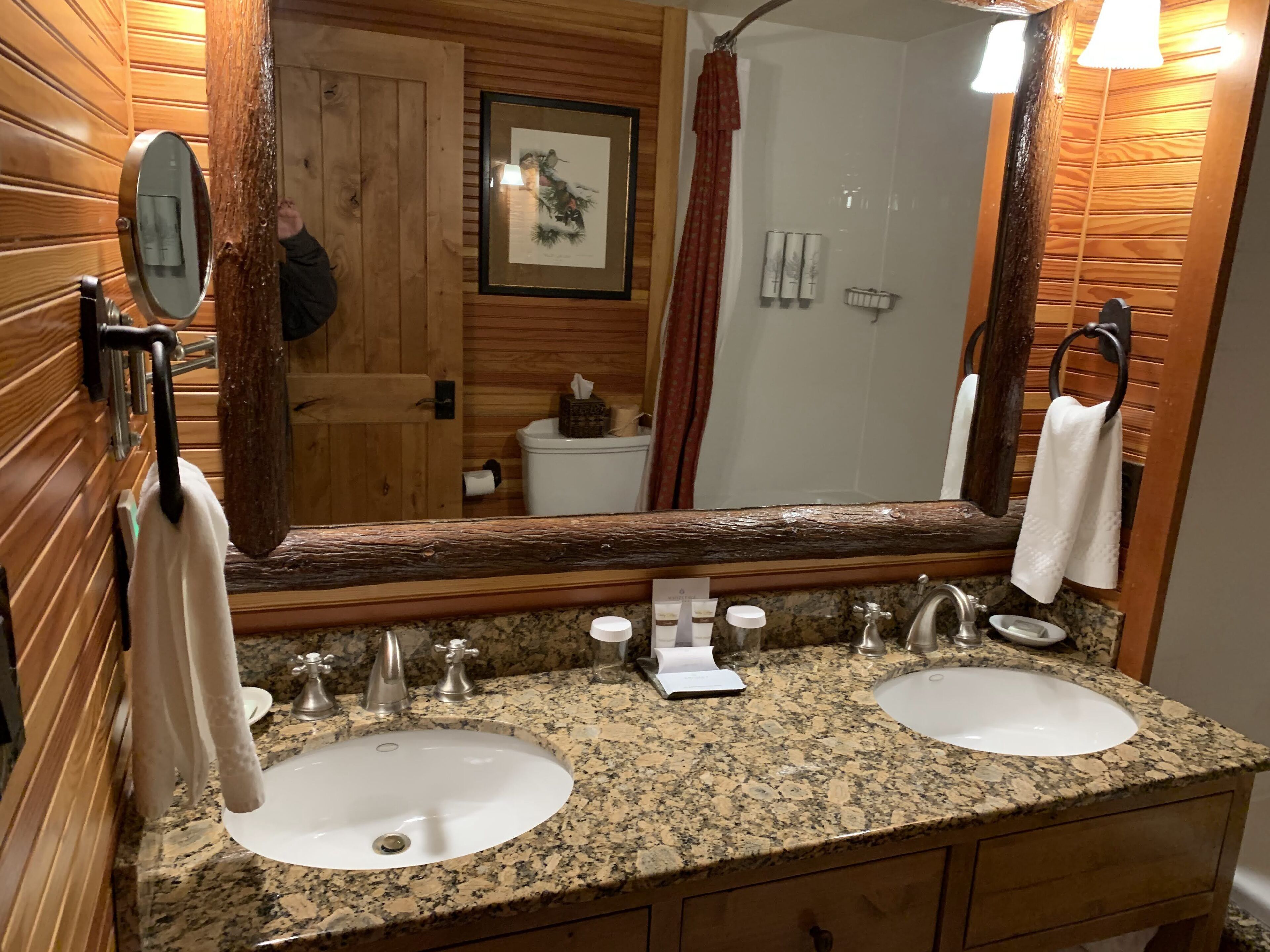 A bathroom with wooden paneling, a double sink, a mirror, and a shower area.