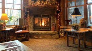 A cozy room with a stone fireplace, wooden furniture, and a view of trees through large windows.