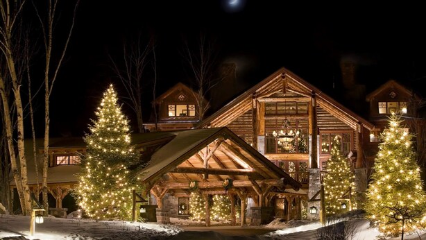 A log cabin with multiple lit Christmas trees in front, a covered porch, and a snow-covered landscape.