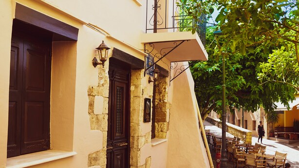 A narrow street with a building featuring a black door, a fire hydrant, and outdoor seating. - Casa Dasyra (Chania)