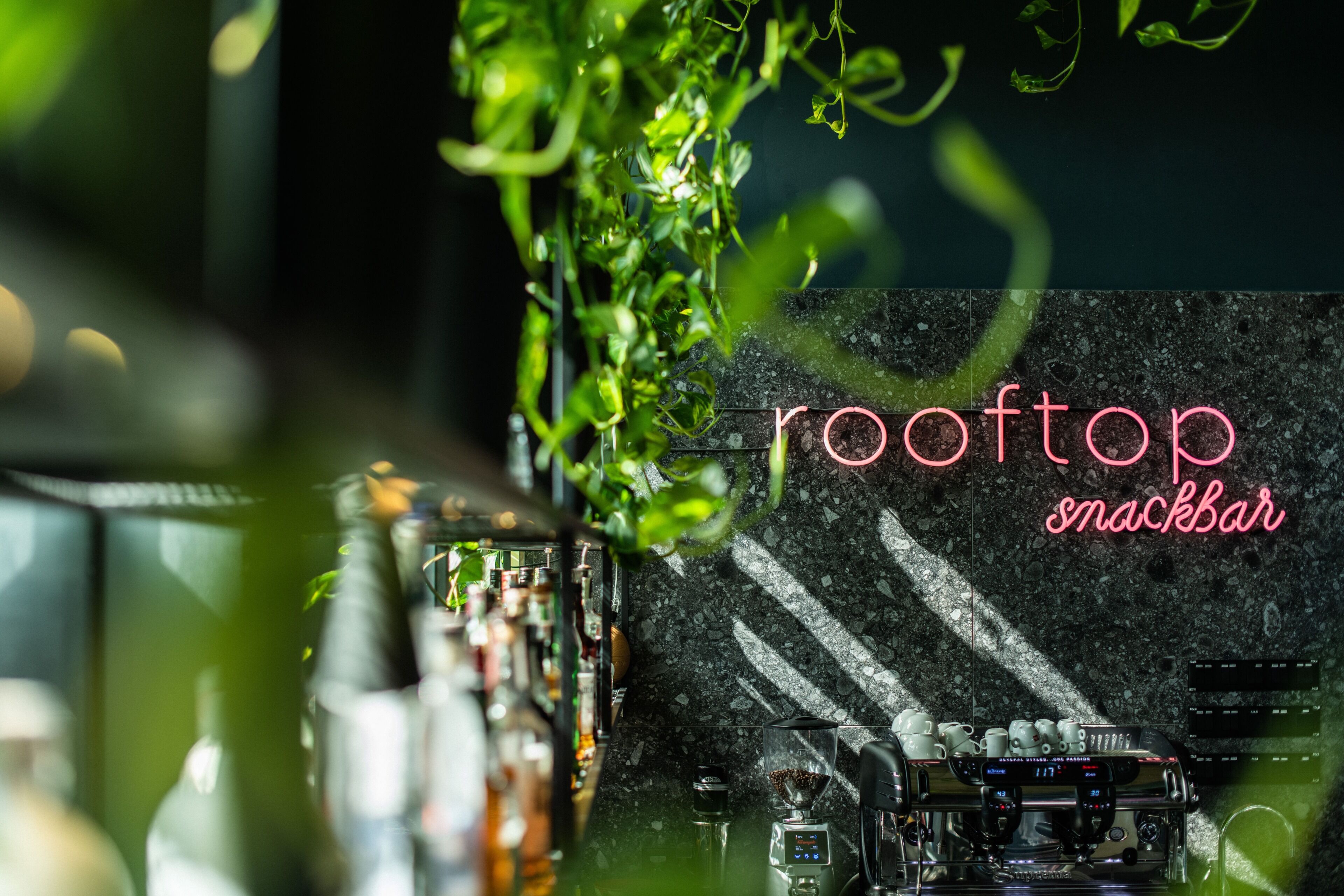 A bar area with a coffee machine, bottles, and a neon sign reading 'rooftop snackbar'.
