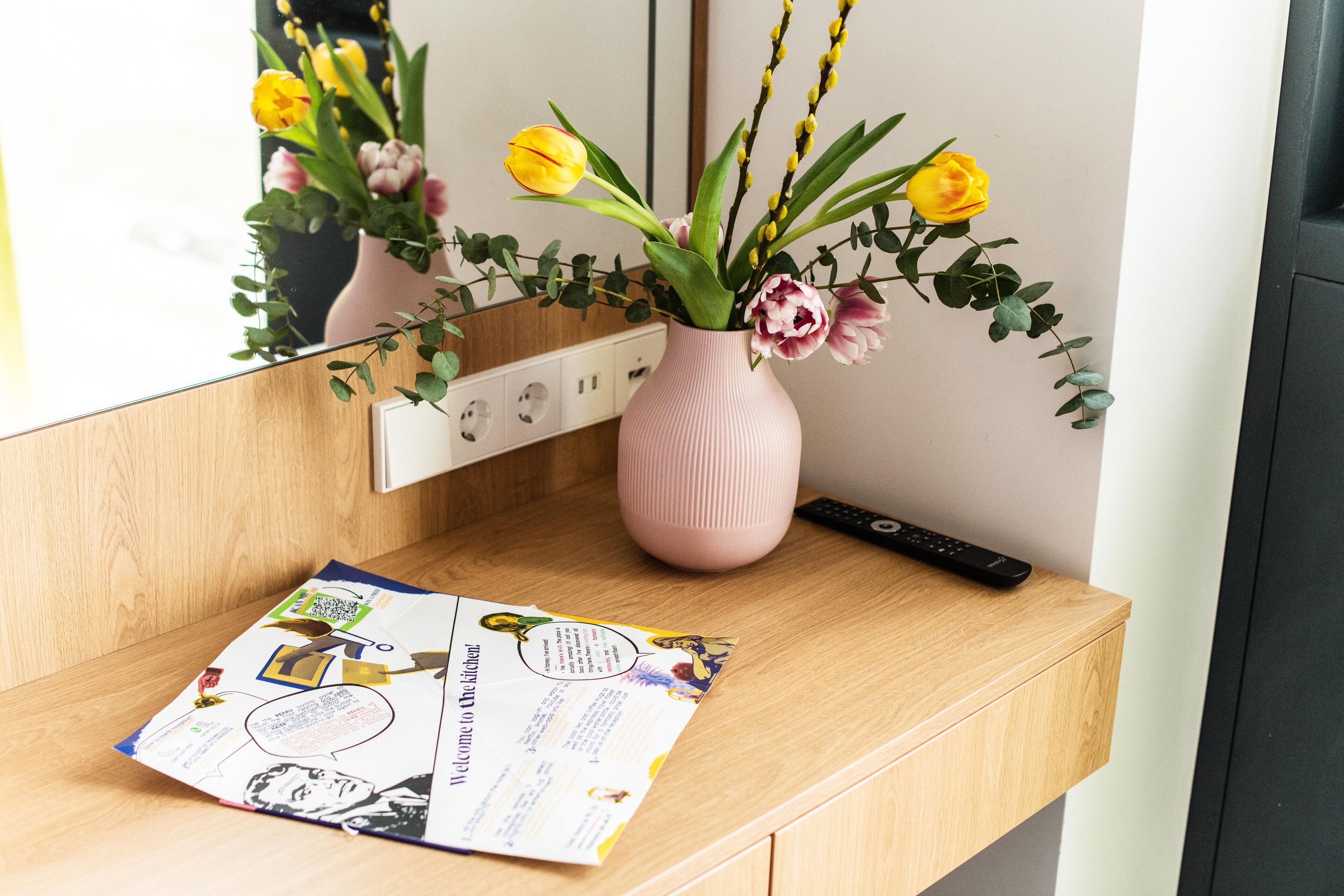 Un bureau en bois sur lequel se trouve un vase rose contenant des fleurs jaunes et une télécommande.
