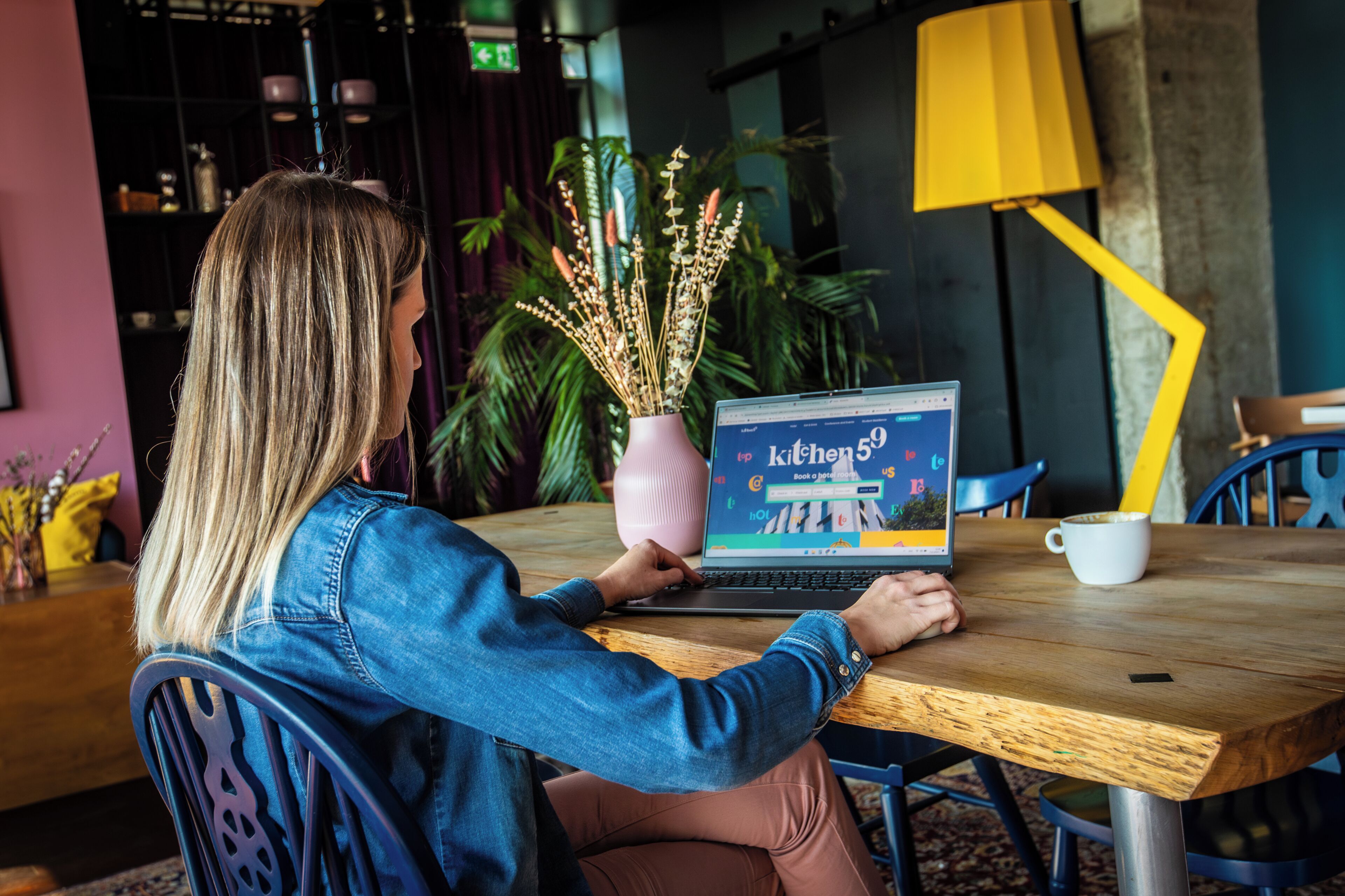 Une personne utilise un ordinateur portable sur une table en bois. On aperçoit en arrière-plan une plante en pot et une lampe jaune.