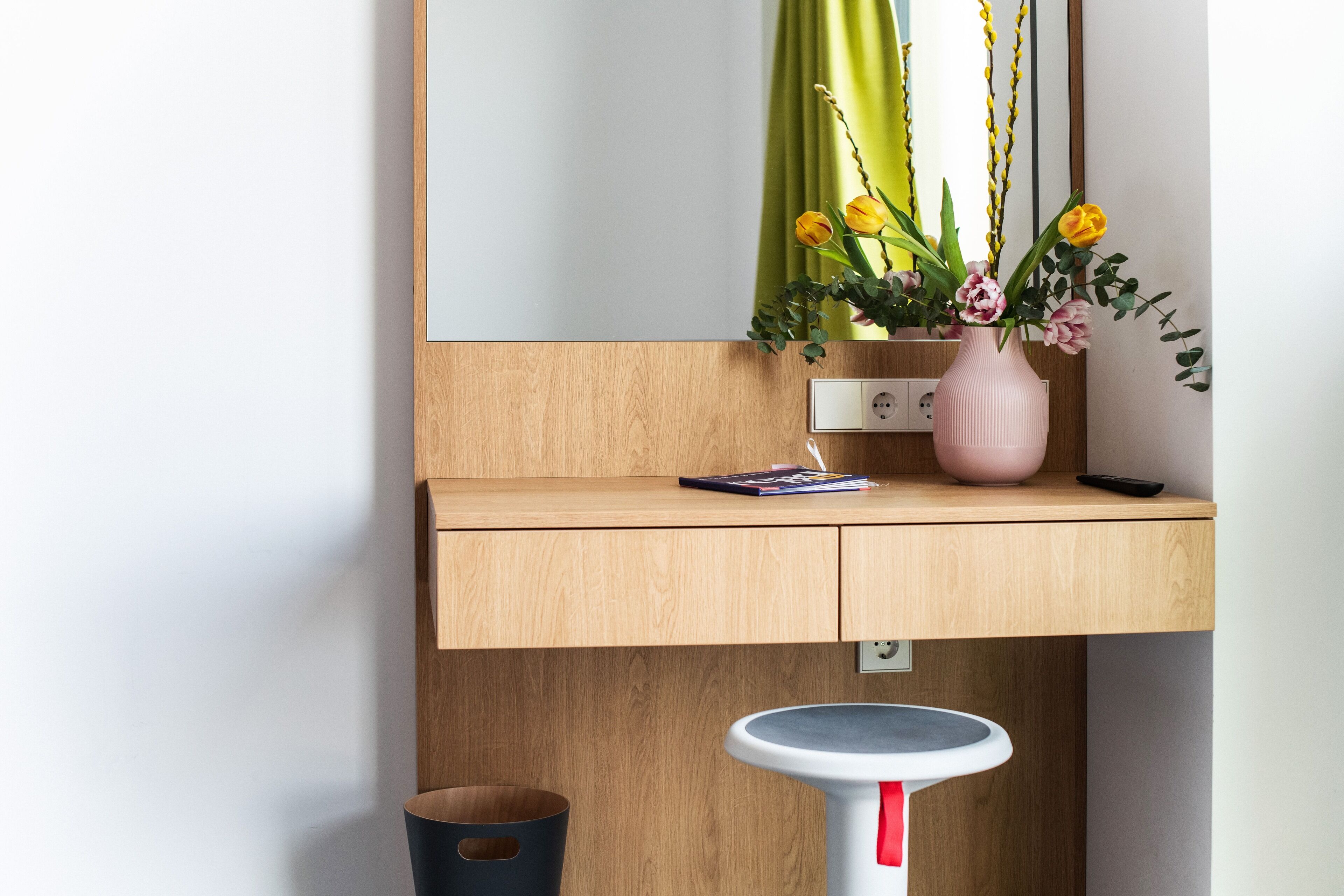 A wooden vanity with a mirror, a pink vase with flowers, a book, and a remote control.