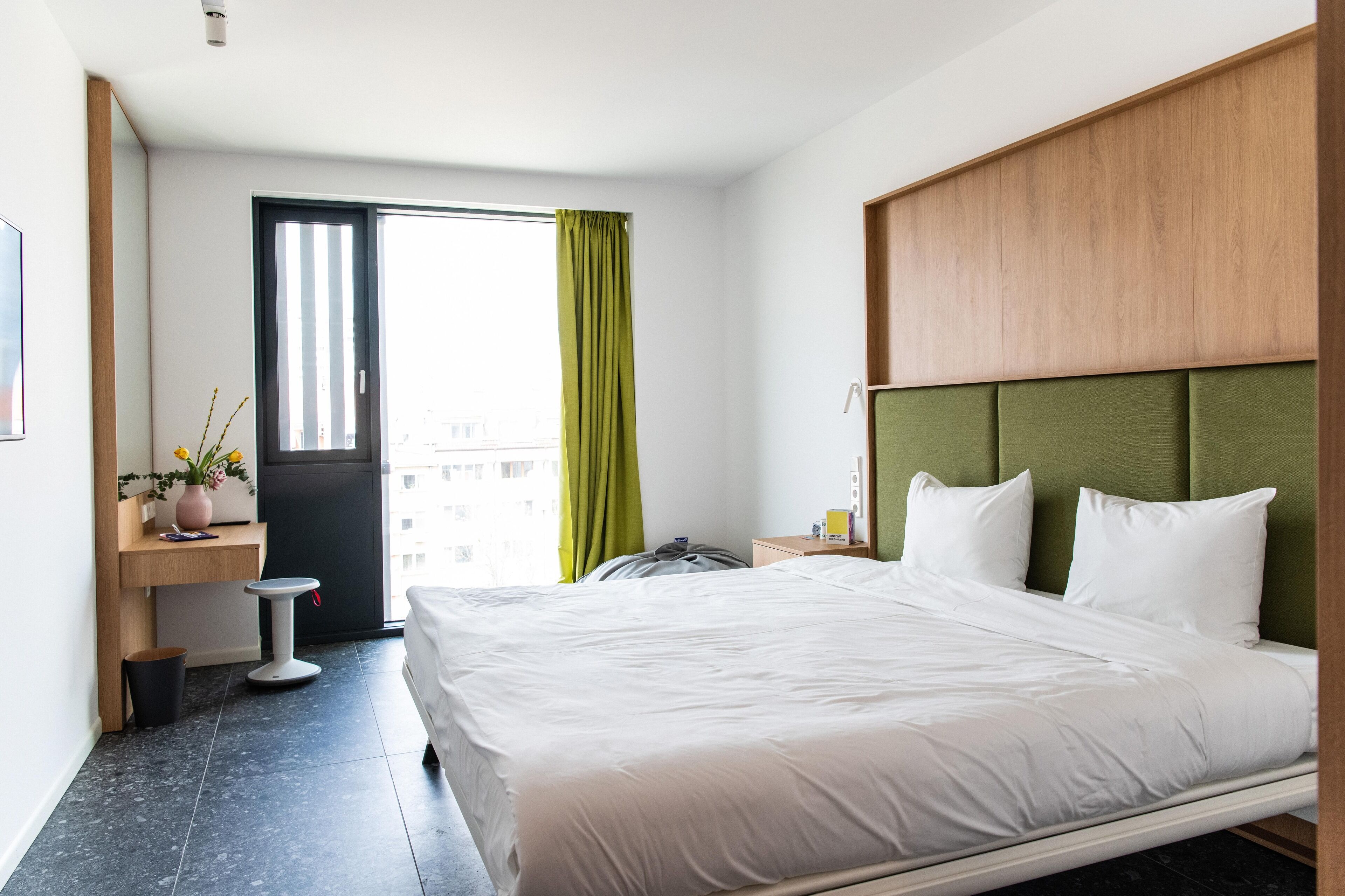A hotel room with a bed, a small desk with a plant, a trash bin, and a window with curtains.