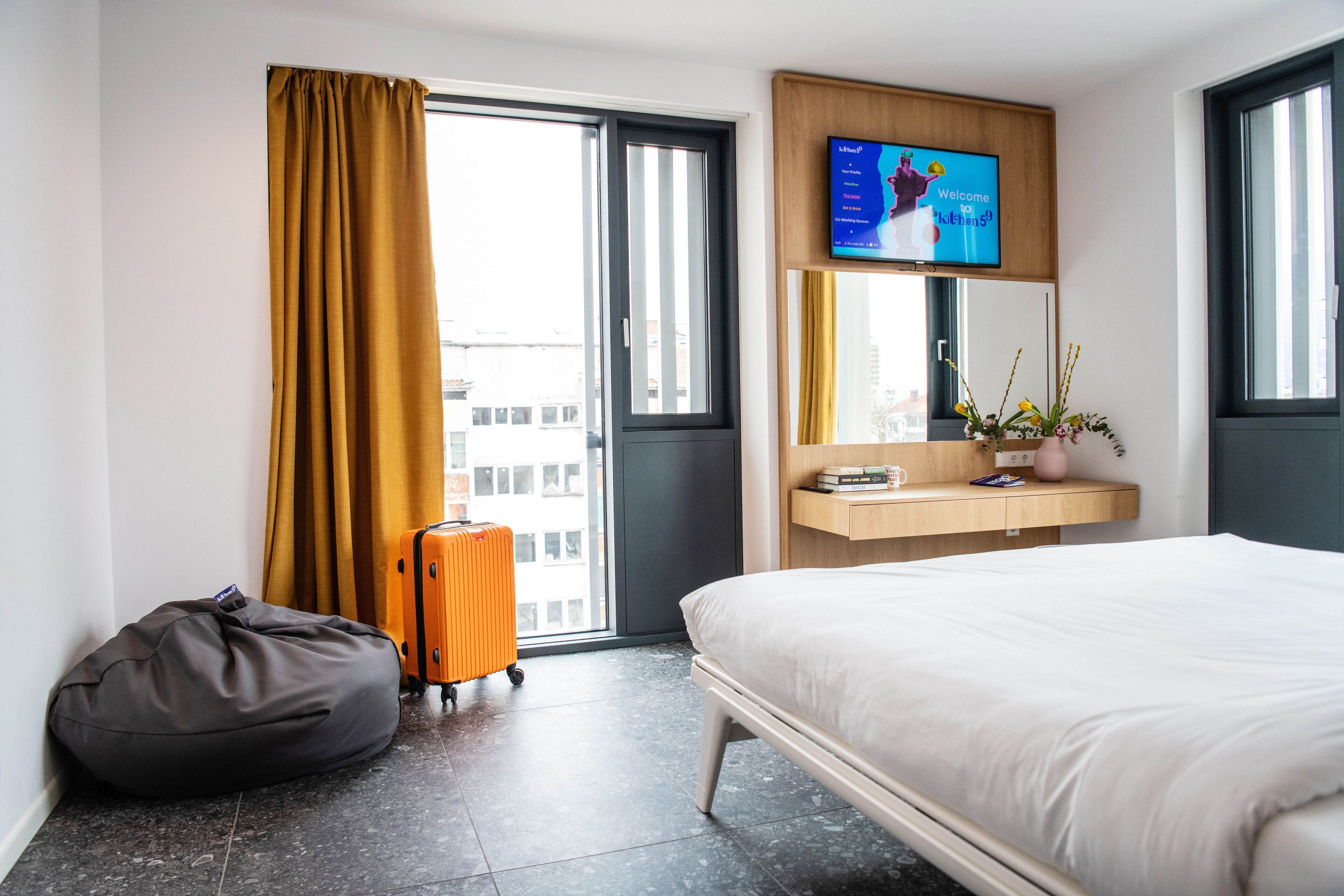 A hotel room with a bed, a desk with a computer monitor, a suitcase, a bean bag, and a window with curtains.