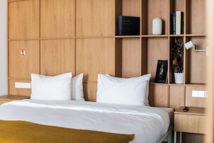 Habitación de hotel con cabecera de madera, cama con ropa blanca, mesita de noche con control remoto y estante con libros y adornos.