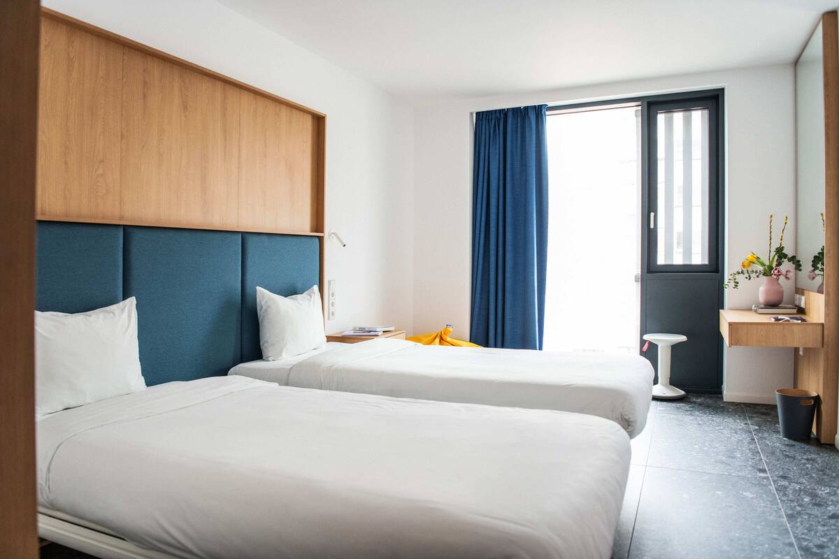 A hotel room with two beds, a wooden headboard, a desk with a chair, a window with blue curtains, and a small plant on the desk.