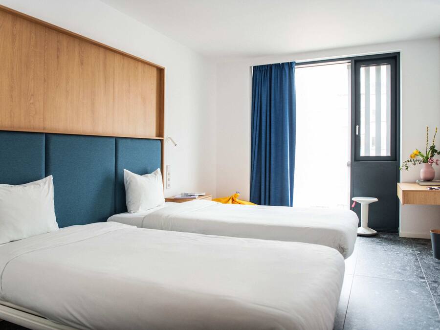 Habitación de hotel con dos camas, cabecera de madera, un escritorio con silla, una ventana con cortinas azules y una planta pequeña sobre el escritorio.
