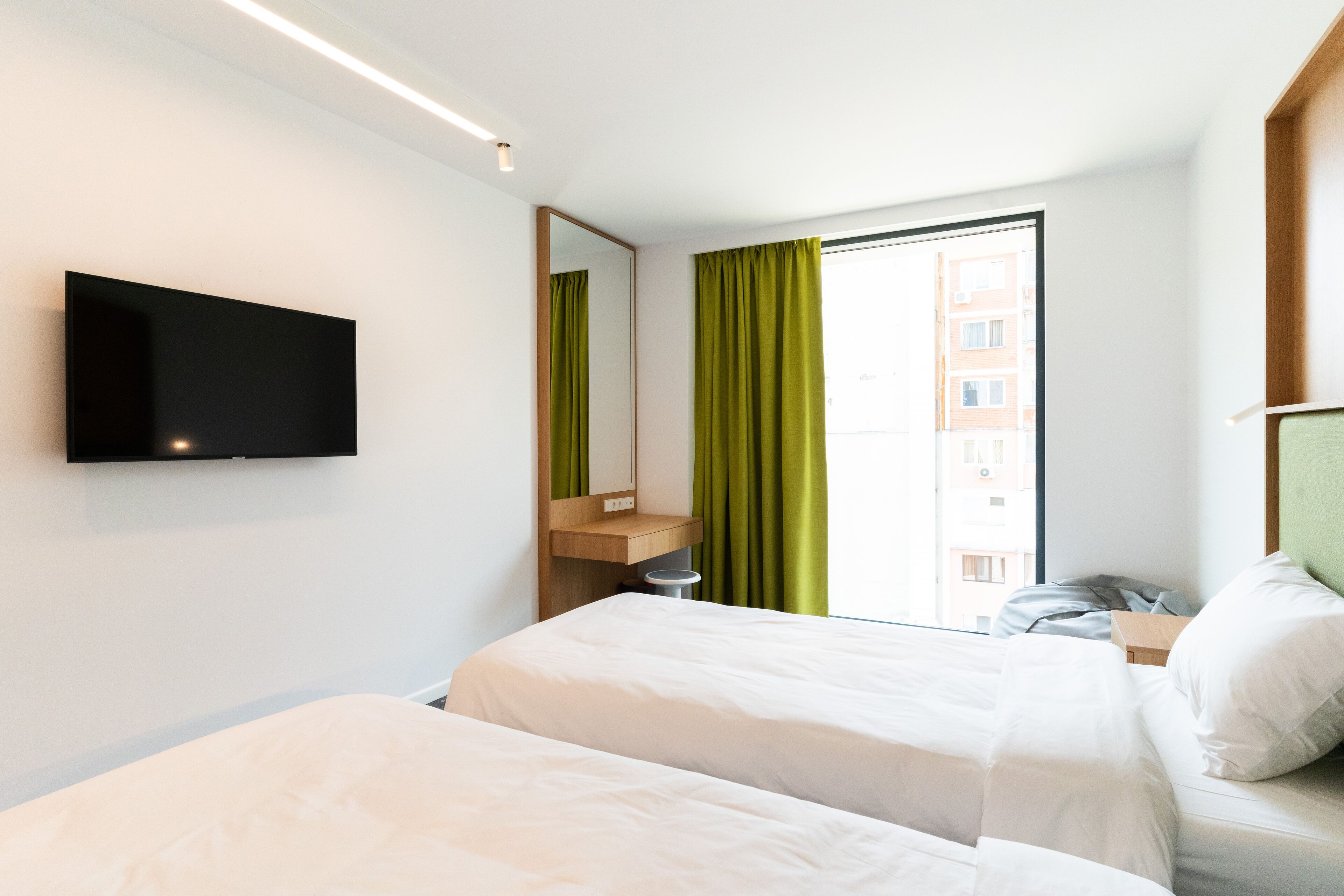 A hotel room with two beds, a TV mounted on the wall, a window with green curtains, and a view of buildings outside.
