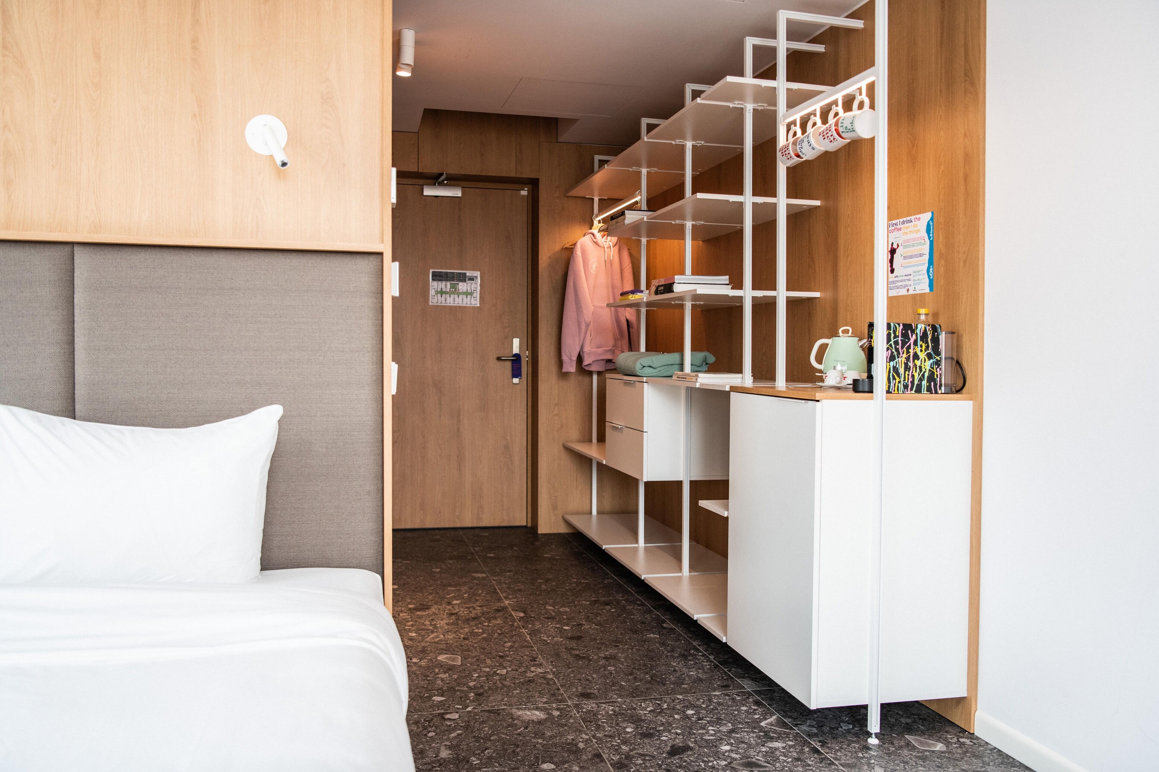 Quarto de hotel com cama, guarda-roupa com prateleiras e pequena mesa com chaleira e chávenas.