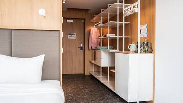 Quarto de hotel com cama, guarda-roupa com prateleiras e pequena mesa com chaleira e chávenas.