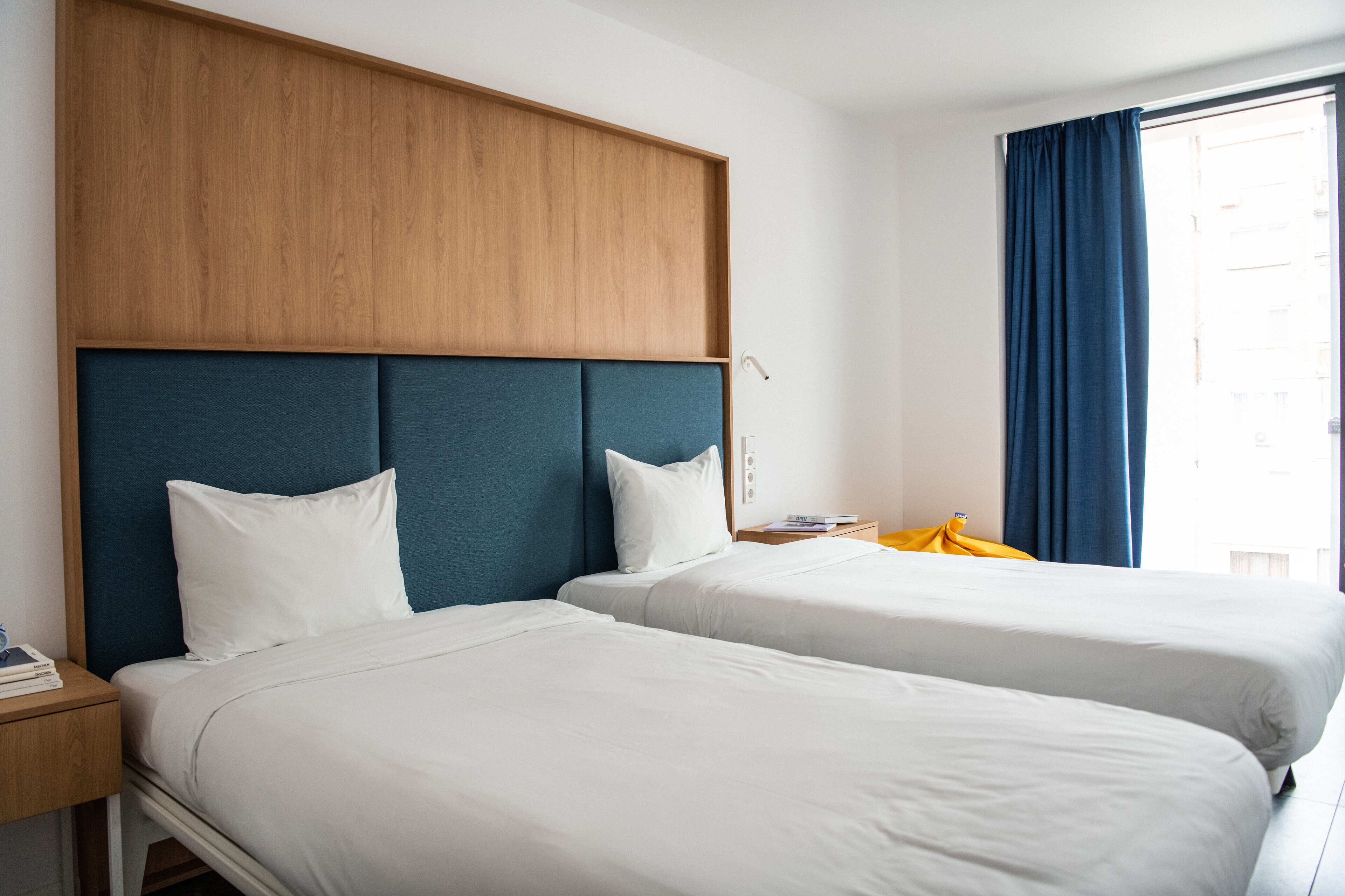 A hotel room with two beds, a wooden headboard, and a large window with blue curtains.