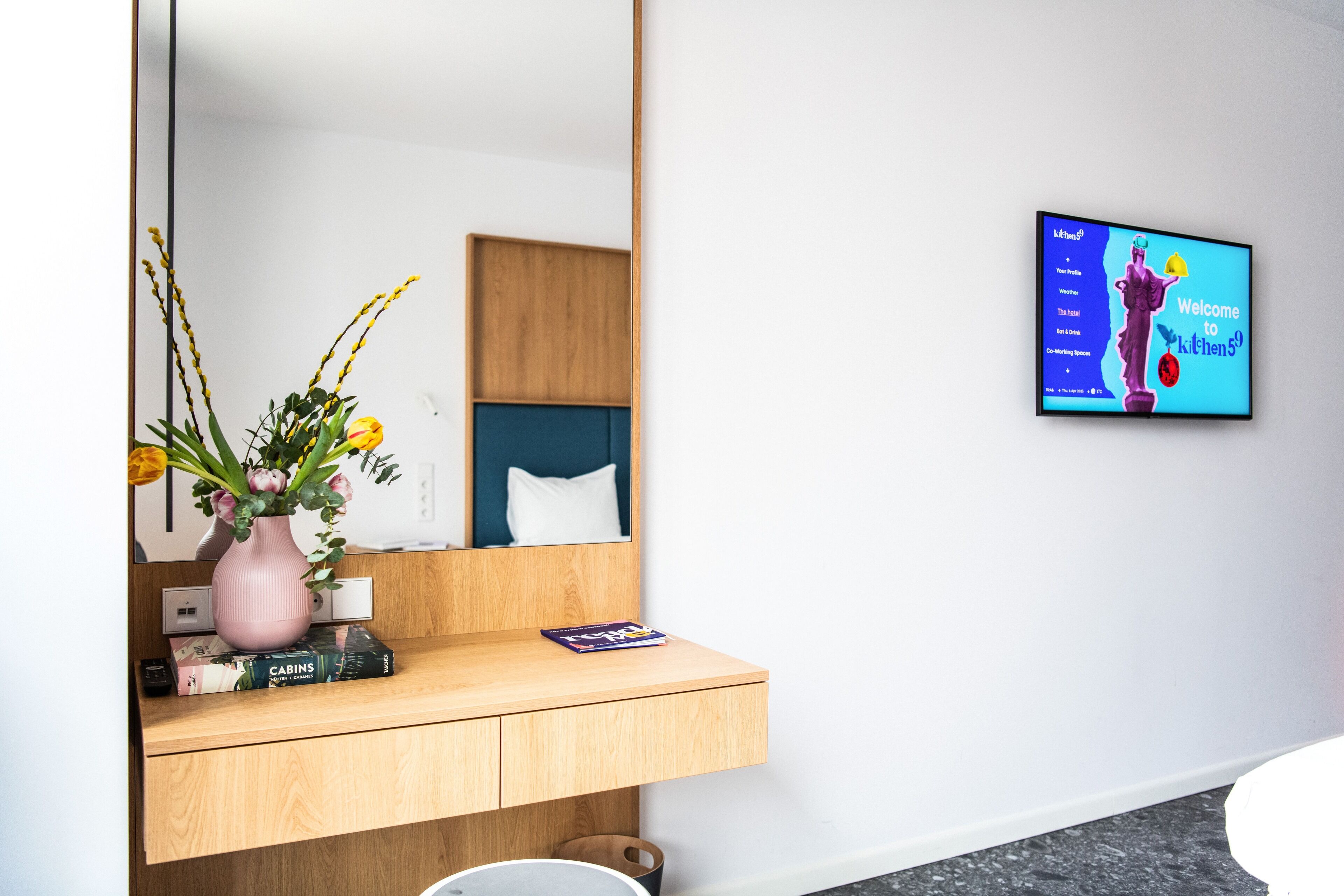 Un baño moderno con tocador de madera, un jarrón rosa con flores y un televisor mostrando un mensaje de bienvenida.
