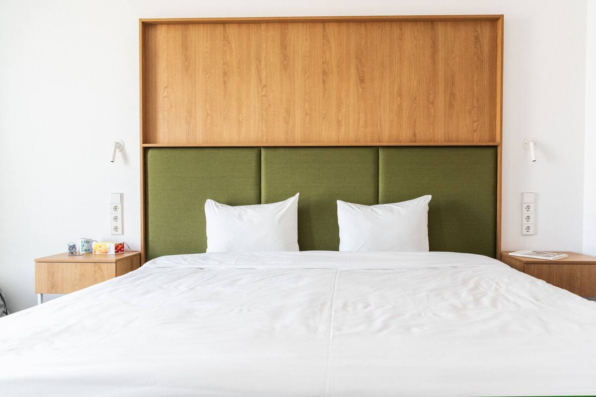 A neatly made bed with a wooden headboard and green upholstered panels, two bedside tables with a small lamp and a book, and a wall with a power outlet.