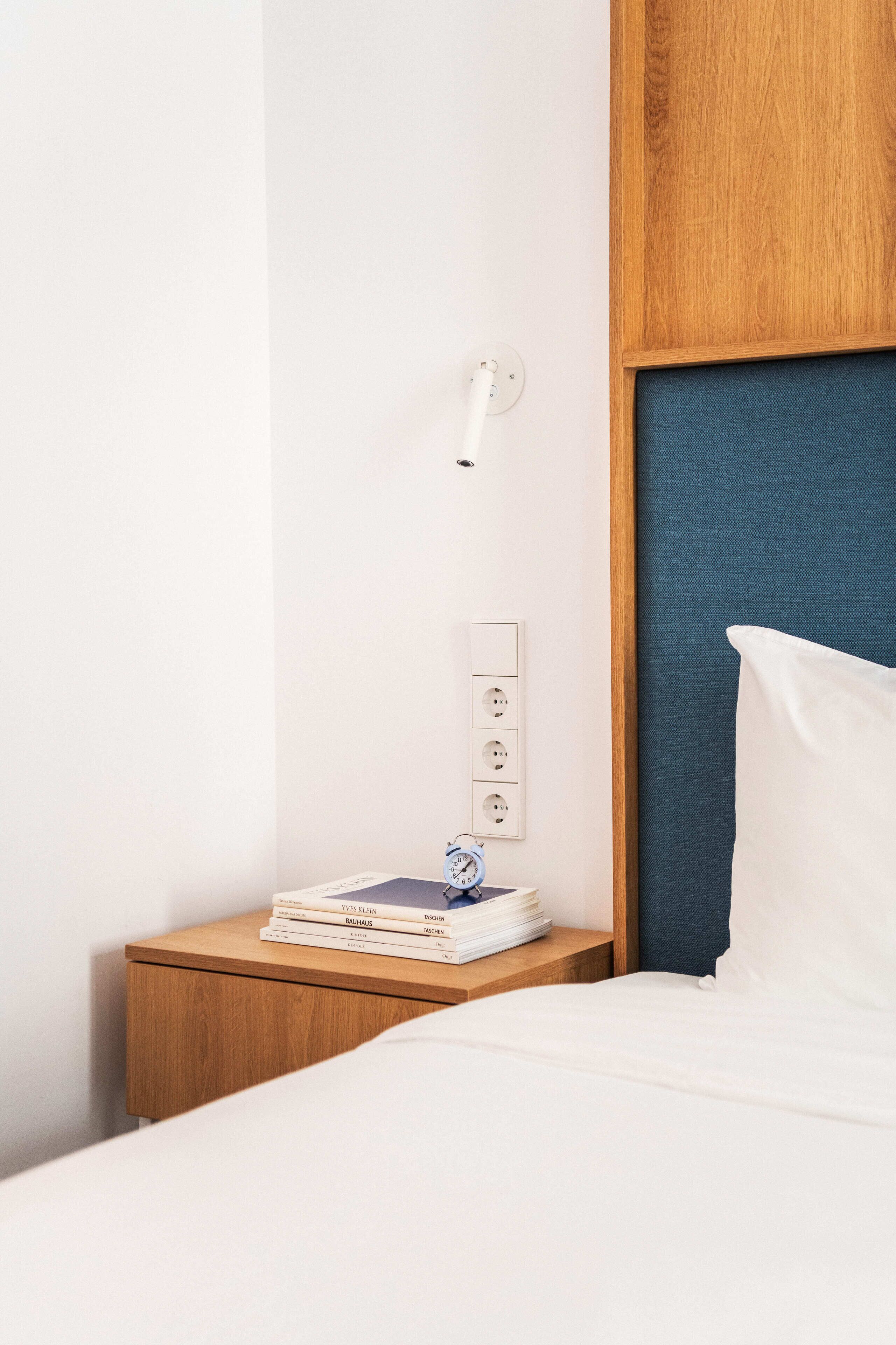 Cama bem arrumada com lençóis brancos, mesa de cabeceira de madeira com uma pilha de livros e uma tomada na parede.