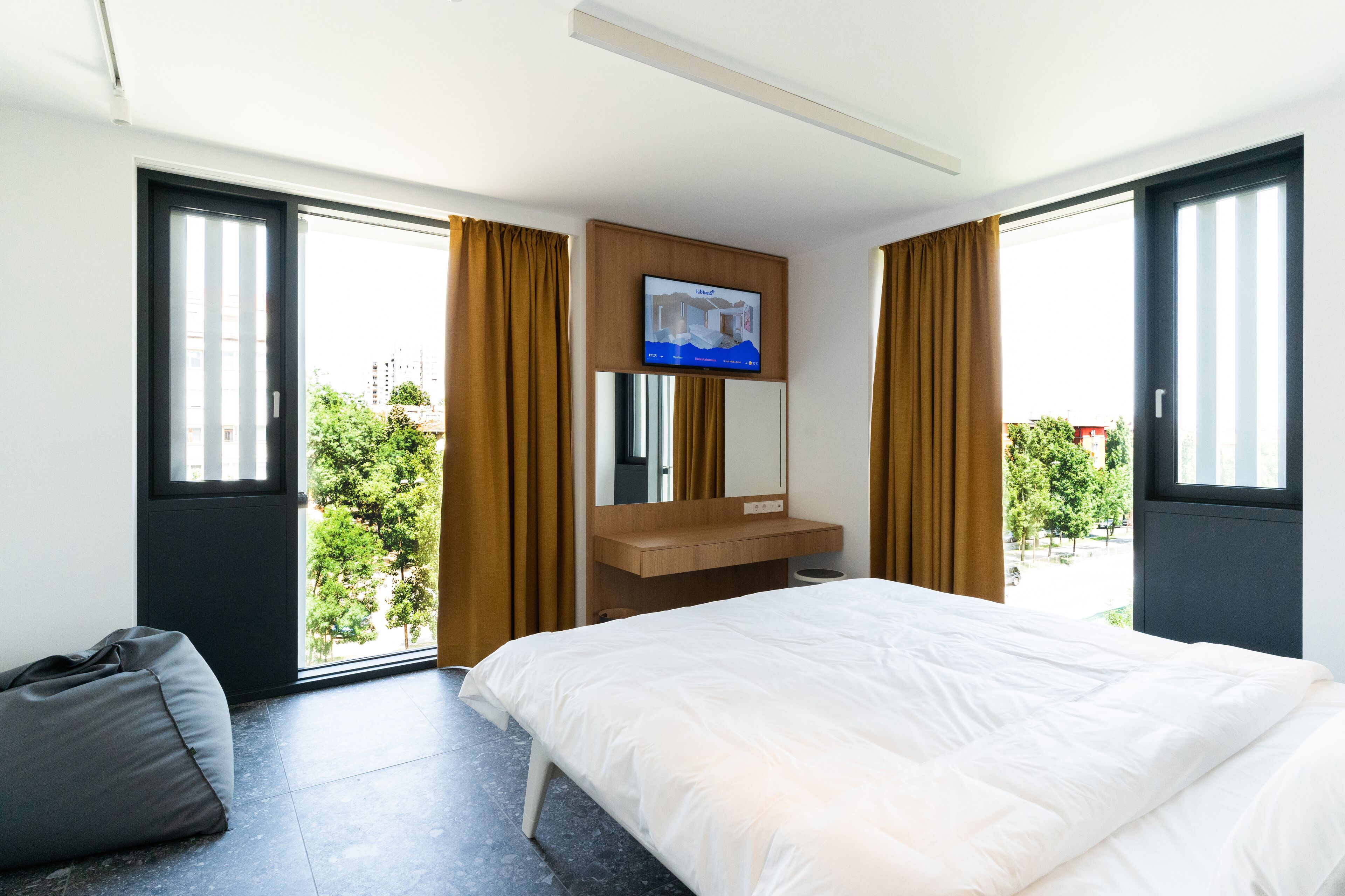 Ein modernes Hotelzimmer mit einem großen Bett, einem an der Wand montierten Fernseher, einem Schminktisch und großen Fenstern mit Vorhängen.