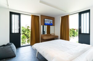 Ein modernes Hotelzimmer mit einem großen Bett, einem an der Wand montierten Fernseher, einem Schminktisch und großen Fenstern mit Vorhängen.