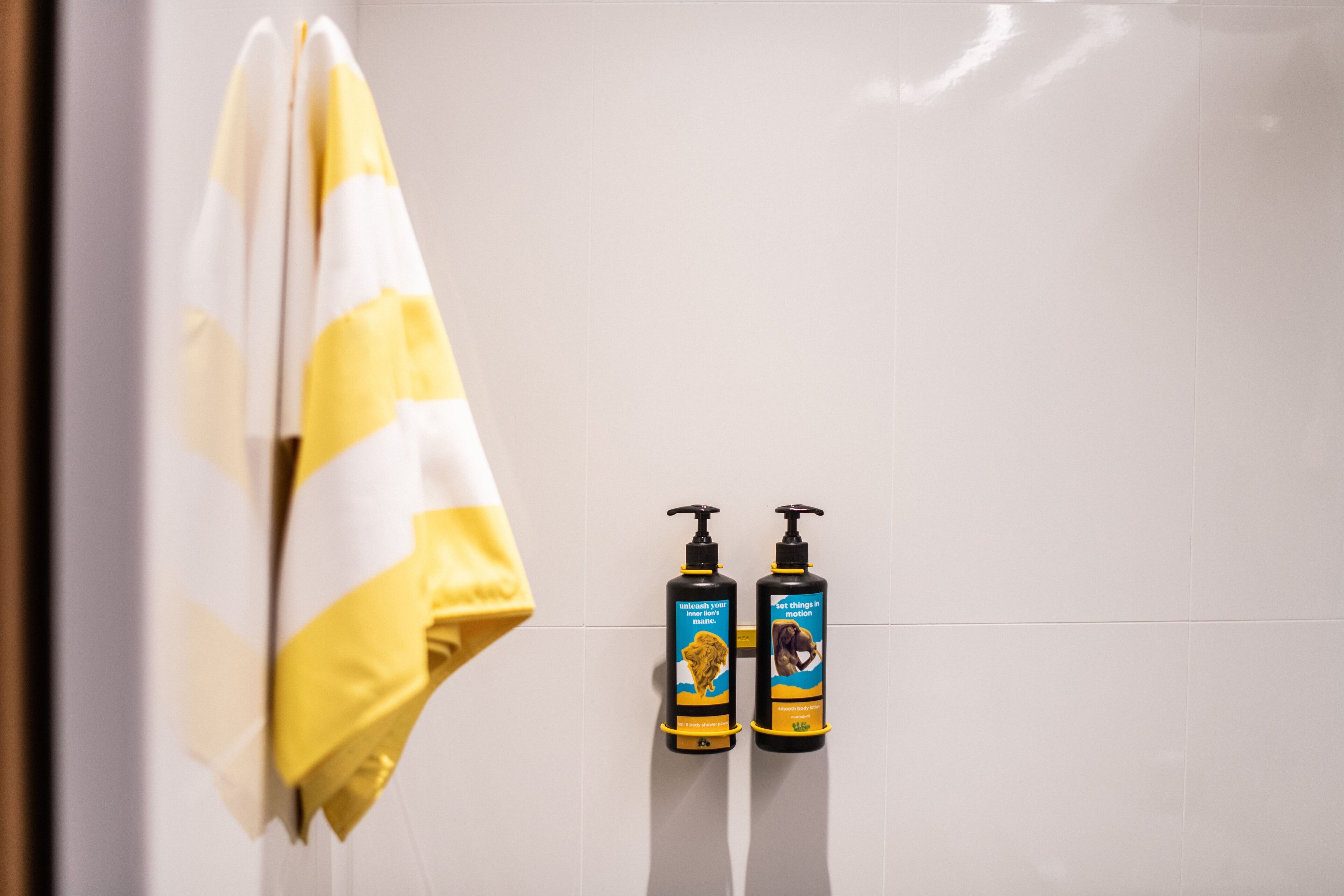 Un baño con una toalla amarilla y blanca a rayas colgada en un perchero, y dos botellas de champú y acondicionador en la pared.