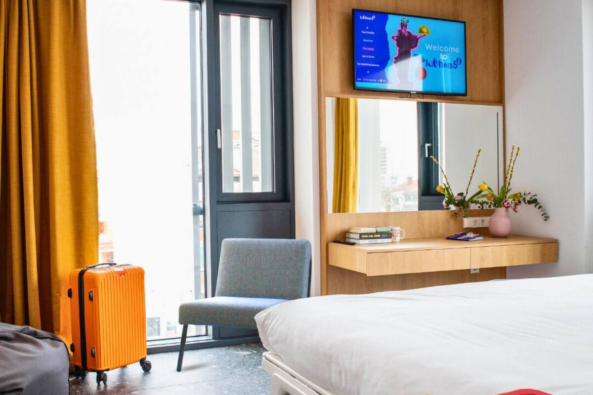 A hotel room with a bed, a desk with a TV, a chair, a suitcase, and a window with curtains.
