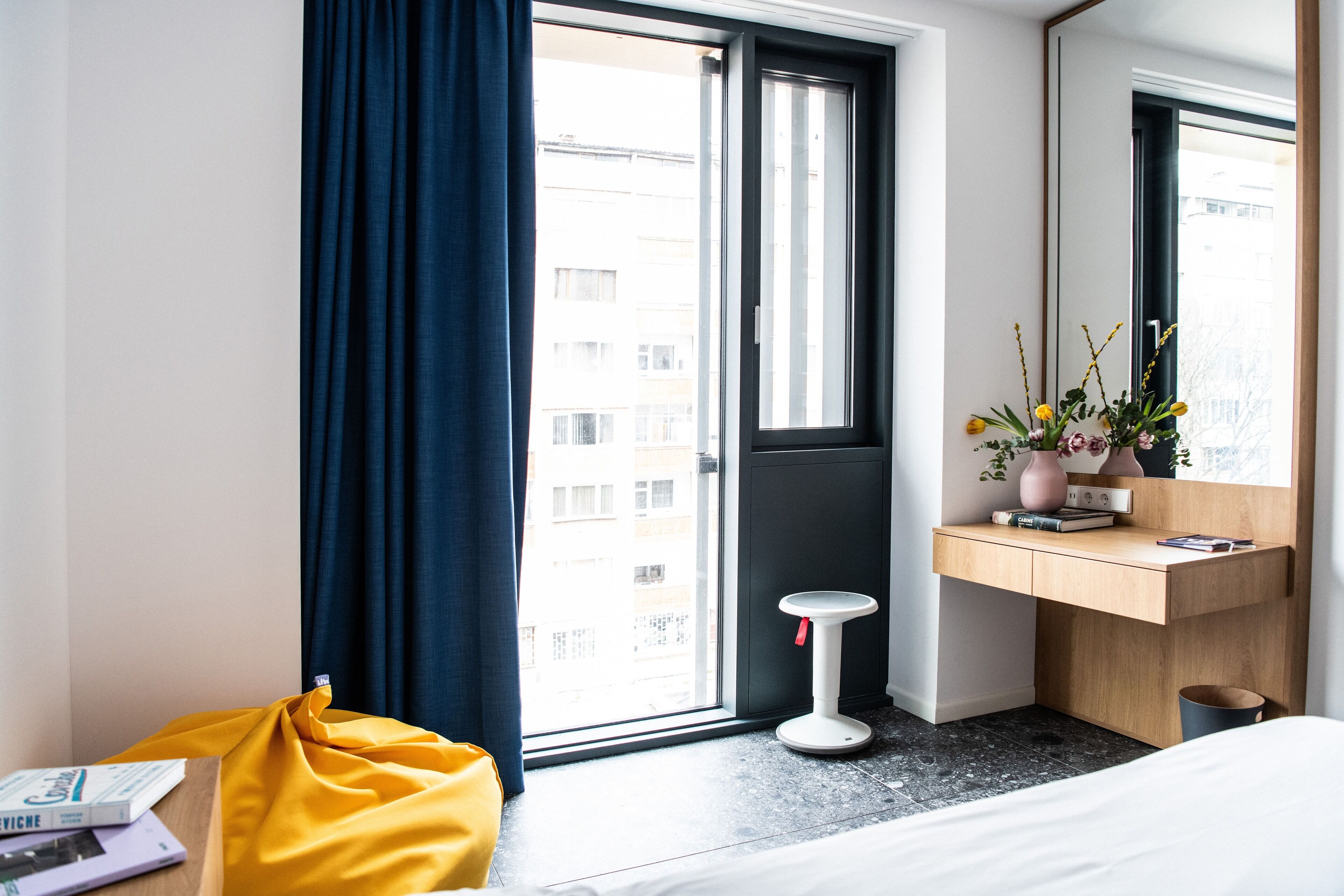 A modern hotel room with a large window, a bedside table with books, a small stool, and a bedside plant.