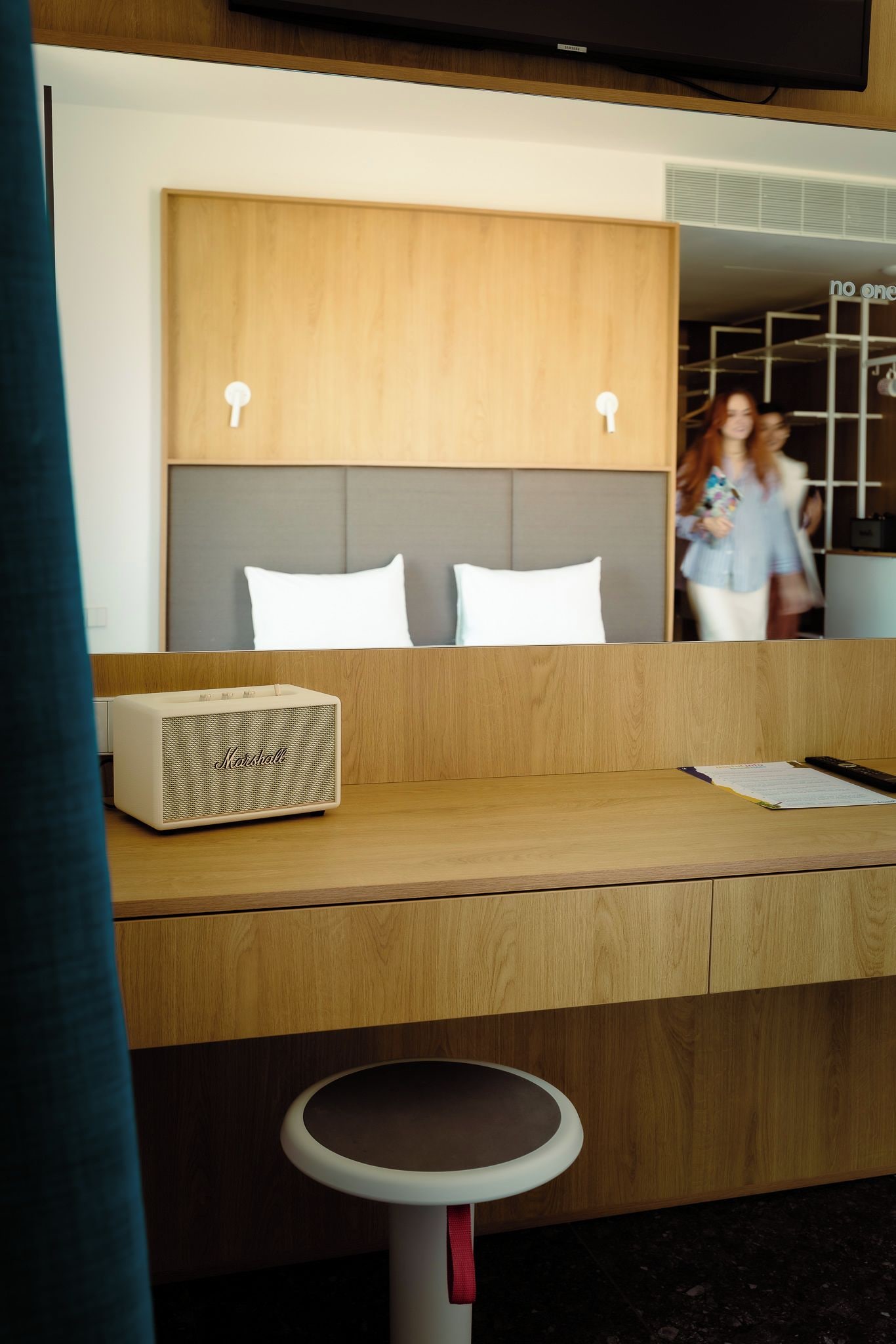 A hotel room with a wooden headboard, a bedside table with a lamp, and a small stool.
